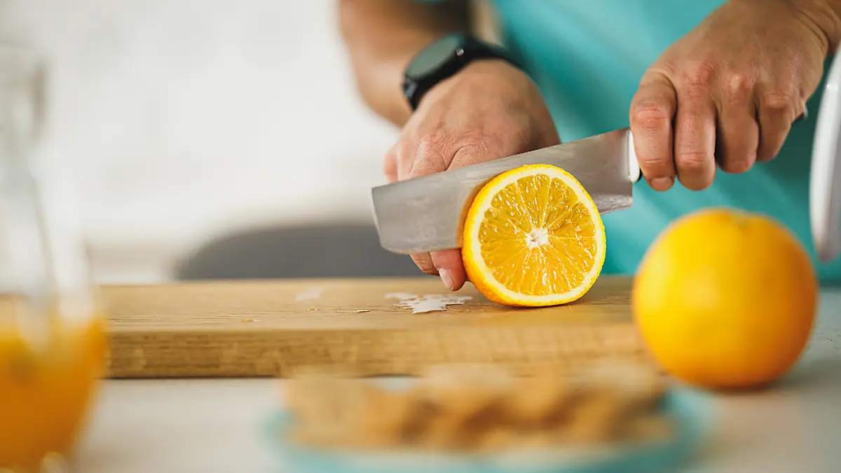 Mãos de adulto cortando uma laranja numa tábua de madeira na cozinha